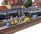 Scène de modélisme ferroviaire avec gare, train et personnages. Des ouvriers chargent des chariots de marchandises. Les bâtiments en arrière-plan créent un décor de petit village.