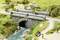 Un petit pont enjambant un ruisseau, entouré de prés et d'arbres. Des personnes se promènent et un cycliste passe à vélo.