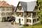 Miniature d'une maison à colombages avec des jardinières fleuries, entourée de personnages dans un village. Une camionnette de livraison est garée à proximité.