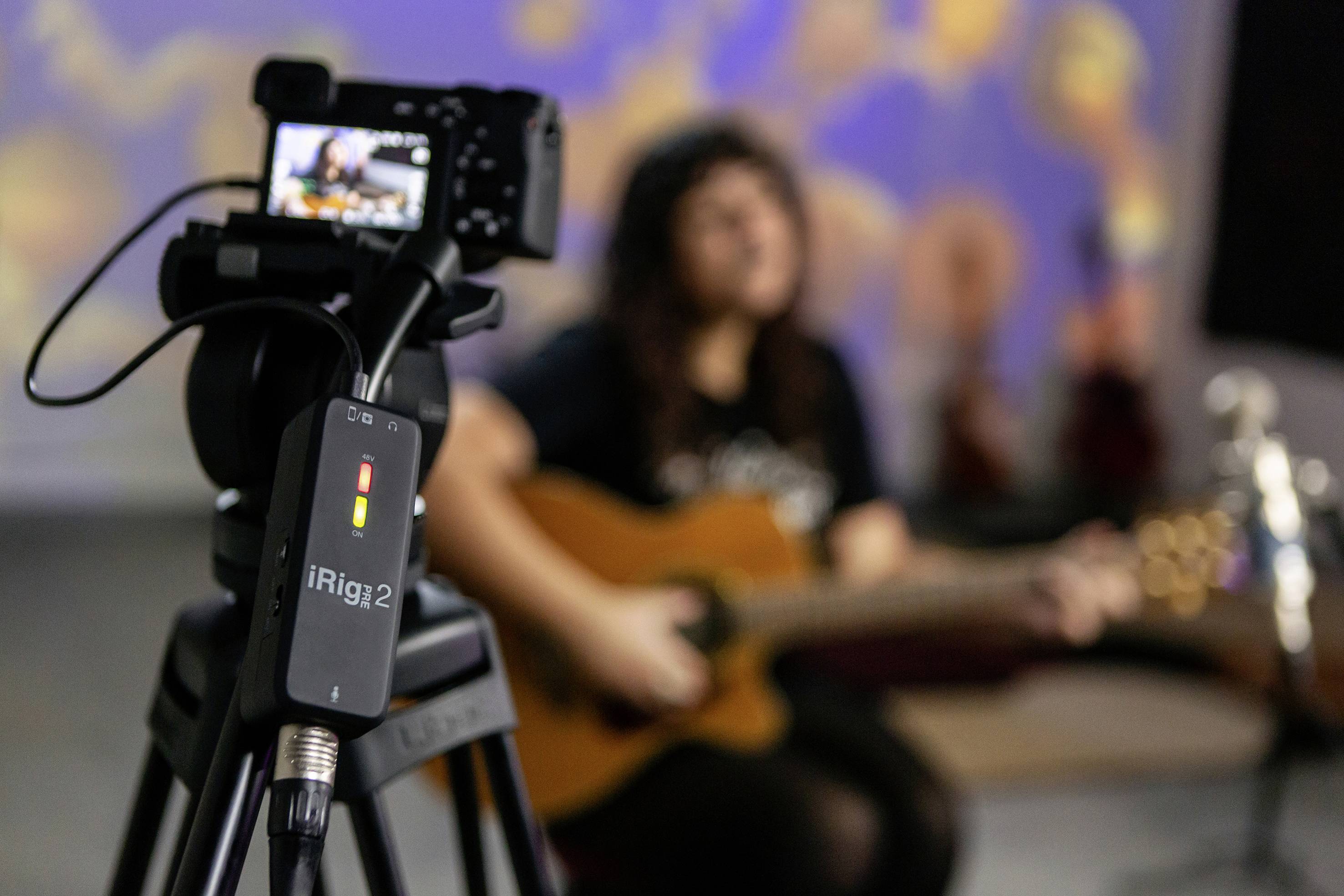 Une caméra au premier plan filme une personne assise qui joue de la guitare. En arrière-plan, la personne est représentée de manière floue.