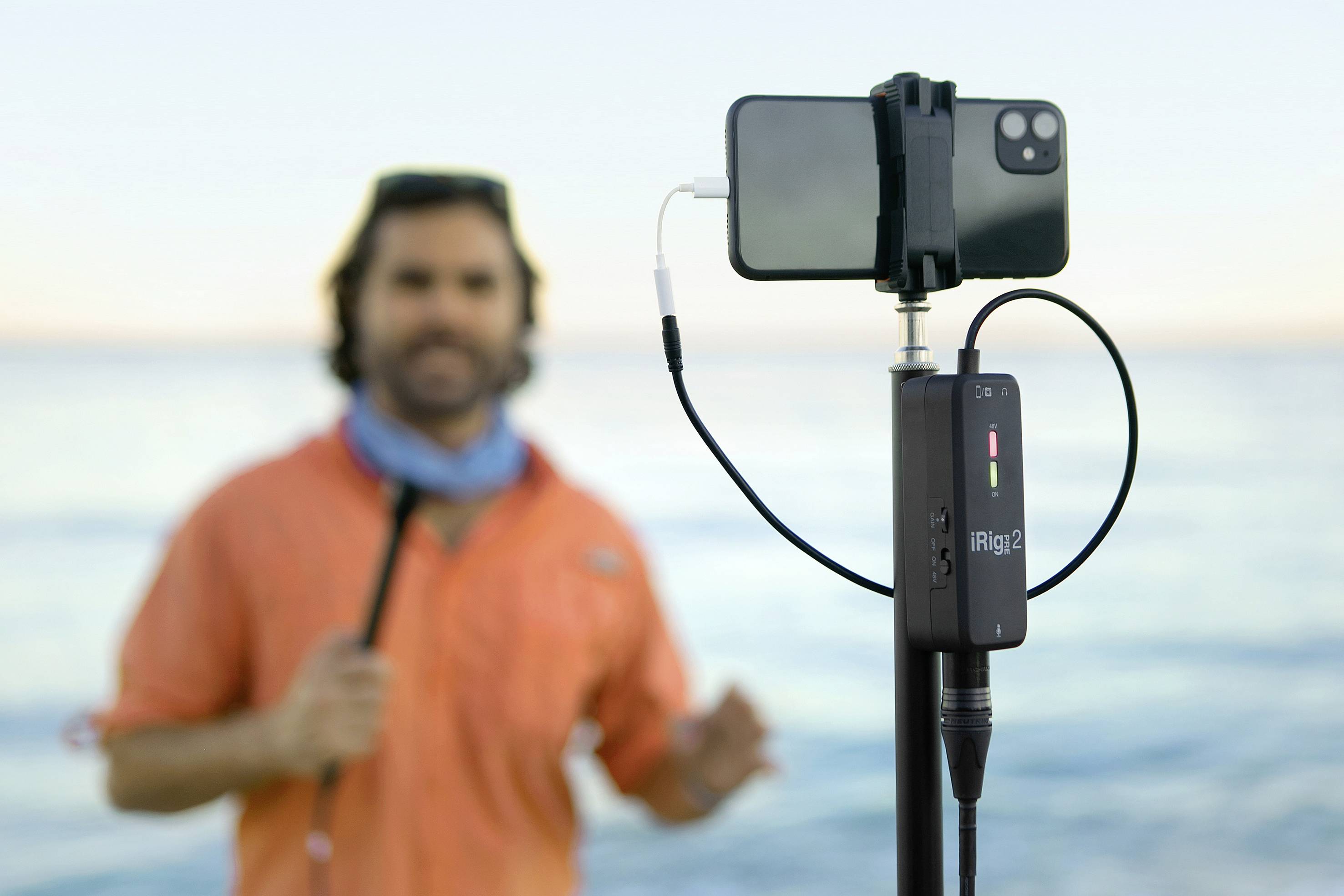 Un homme vêtu de vêtements orange se tient près de la mer et est filmé avec un smartphone monté sur un trépied. Un interface audio est visible au premier plan.