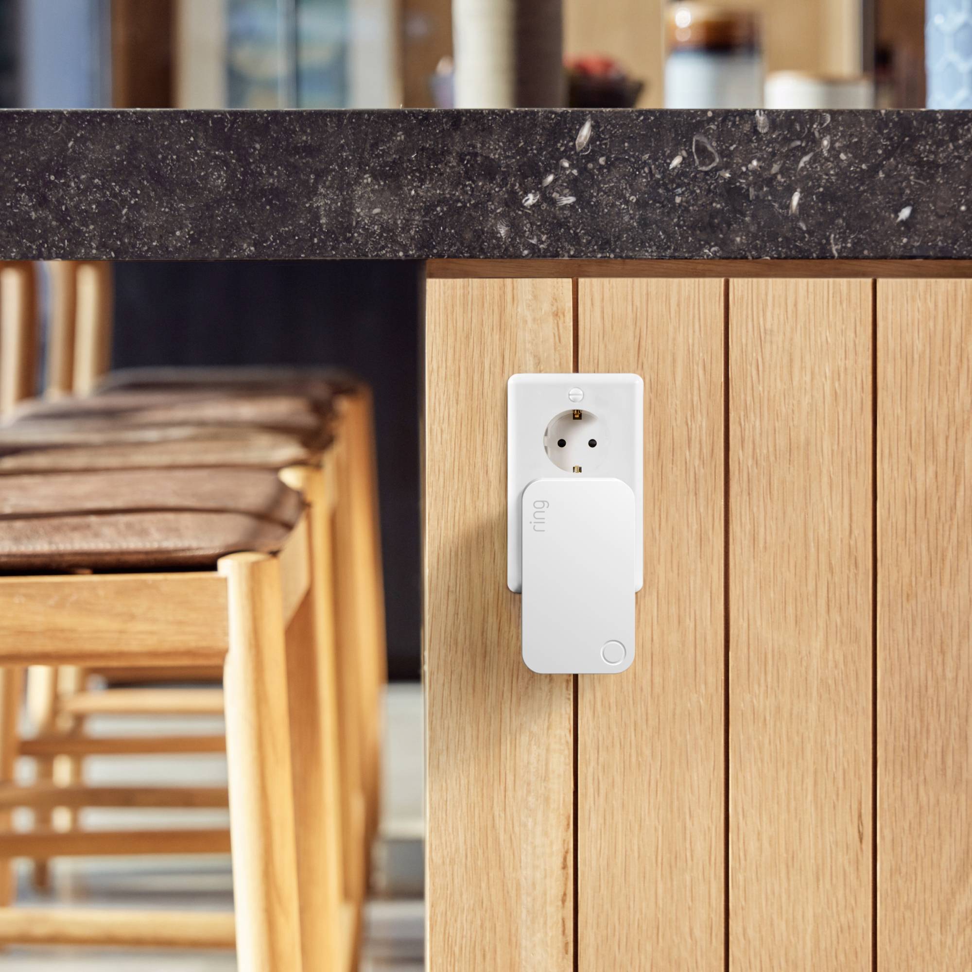 Une prise connectée est installée sur le mur de la cuisine. En arrière-plan, des chaises modernes en bois et une table sont floues.