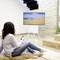 Une femme est assise sur un tapis dans un salon et regarde la télévision. L'écran montre un paysage de plage. Elle tient une télécommande dans sa main.