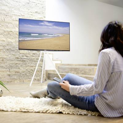 Une personne est assise dans un salon moderne et regarde un grand téléviseur à écran plat sur lequel est affiché une image de plage.