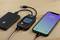 Un smartphone affiche l'heure '8:42' et est connecté à un testeur de performance USB, lui-même branché à une batterie externe.