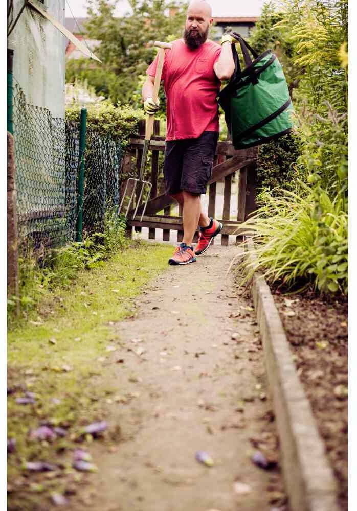 Un homme portant un t-shirt rouge et un pantalon noir transporte un grand sac vert sur un chemin de jardin.