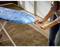 Une personne tend une housse bleue sur une planche à repasser. La planche à repasser est posée sur un tapis dans une pièce au sol en bois.