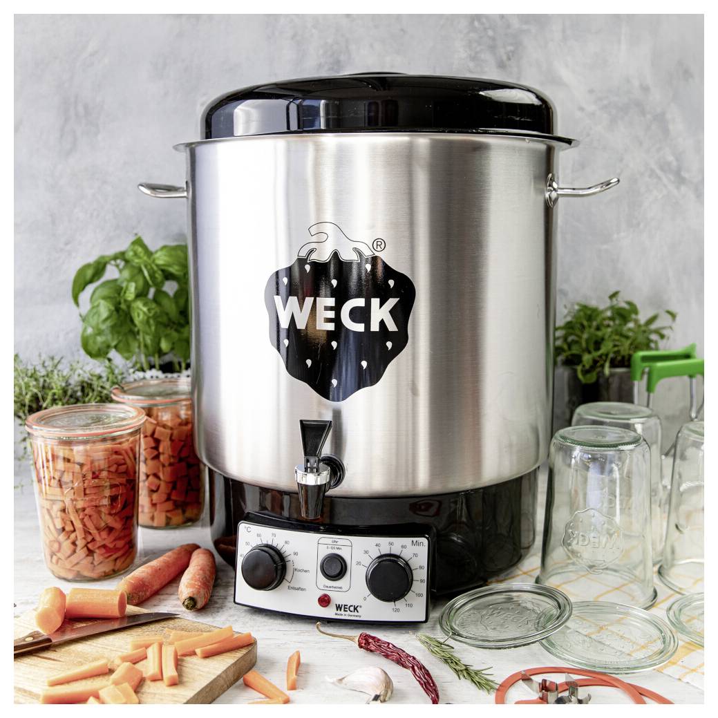 Une casserole de conserves Weck en acier inoxydable avec un cadran de température est posée sur un comptoir de cuisine, à côté de bocaux, de carottes tranchées et d'herbes fraîches.