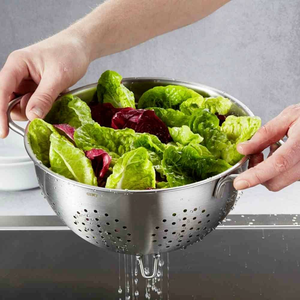 Deux mains tiennent un tamis rempli de laitue fraîchement lavée au-dessus d'un évier. L'eau goutte de la salade.