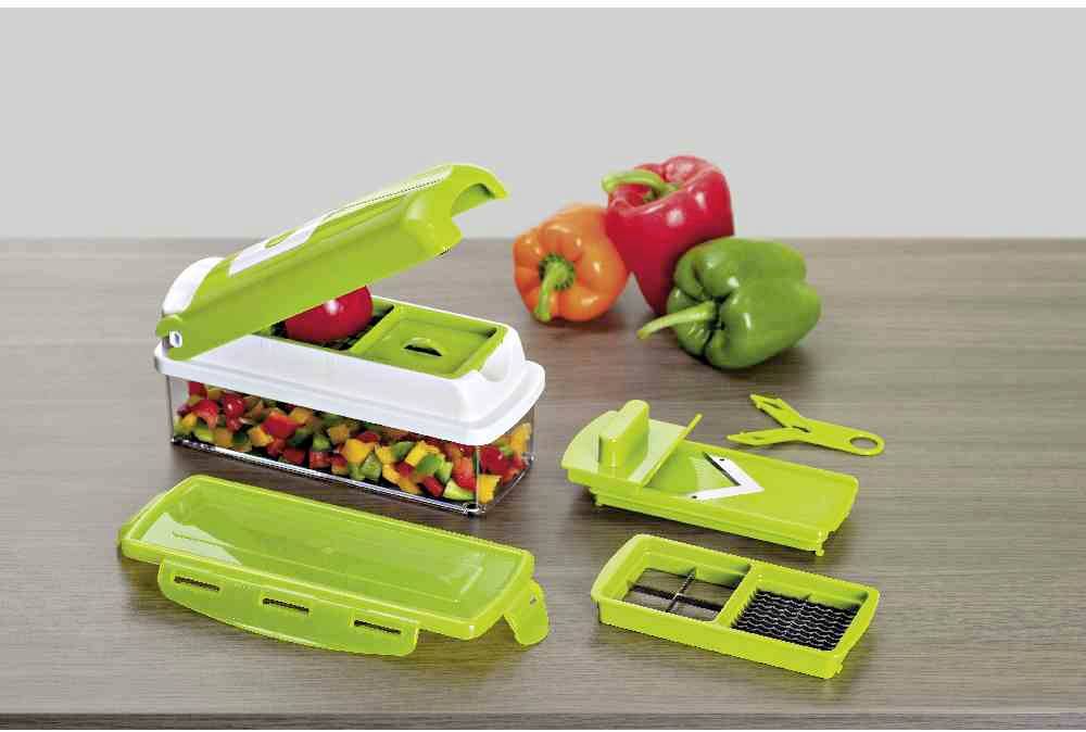 Un coupe-légumes posé sur une table avec plusieurs accessoires et trois poivrons en rouge, jaune et vert. Le récipient est rempli de légumes coupés.
