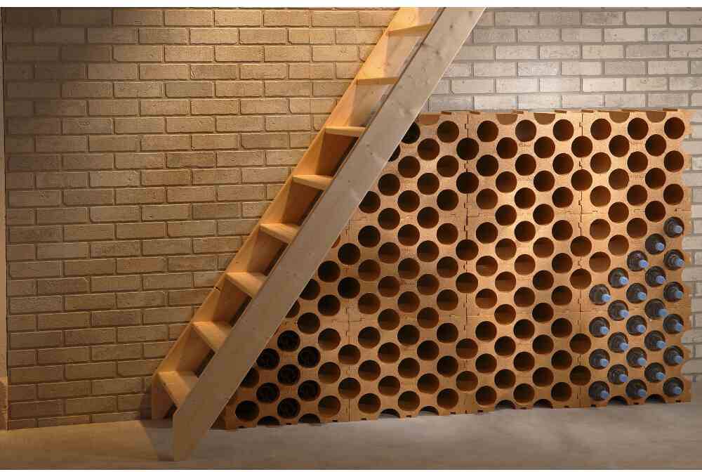 Cave à vin sous un escalier en bois avec un mur de briques, remplie de bouteilles sur des étagères rondes ouvertes. Une cave à vin aux allures chaleureuses et accueillantes.