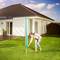 Une femme installe un étendoir à linge dans le jardin. À l'arrière-plan, on peut voir une maison blanche avec une terrasse et des plantes.