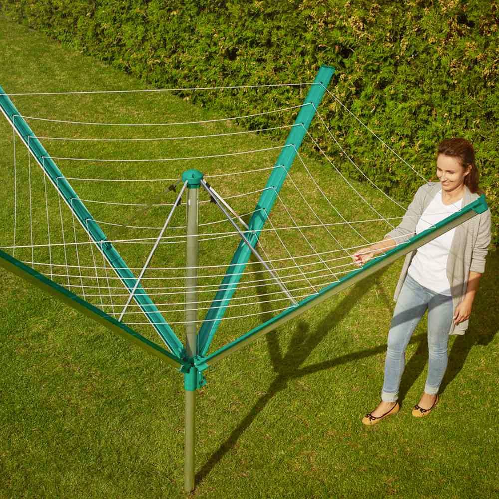 Une femme se tient à côté d'un parasol vert dans le jardin. Elle regarde les cordes à linge, devant une haie verte. Il fait soleil dehors.