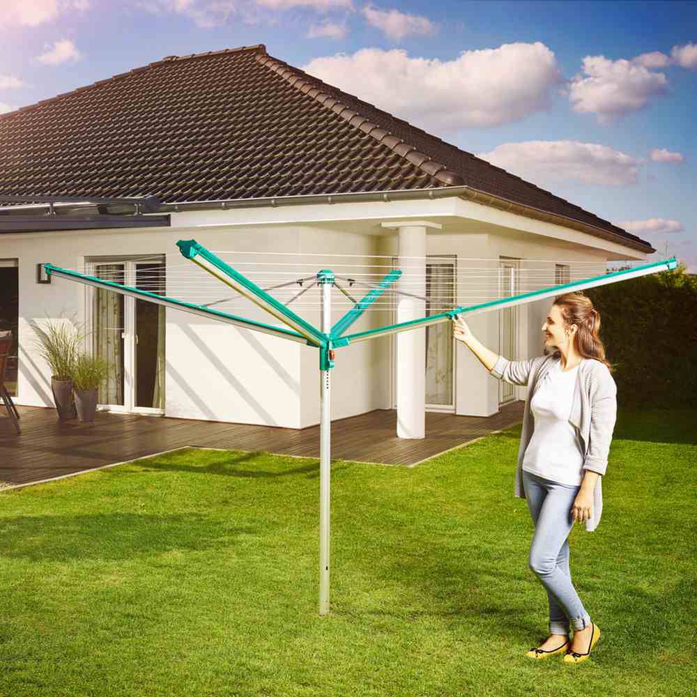 Une femme se tient dans le jardin à côté d'un étendoir à linge pliable. À l'arrière-plan, on peut voir une maison blanche avec un toit brun.