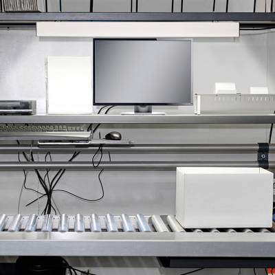 Un poste d'emballage avec un écran, un clavier et un tapis roulant. Un carton flotte sur le tapis roulant, prêt à être traité.