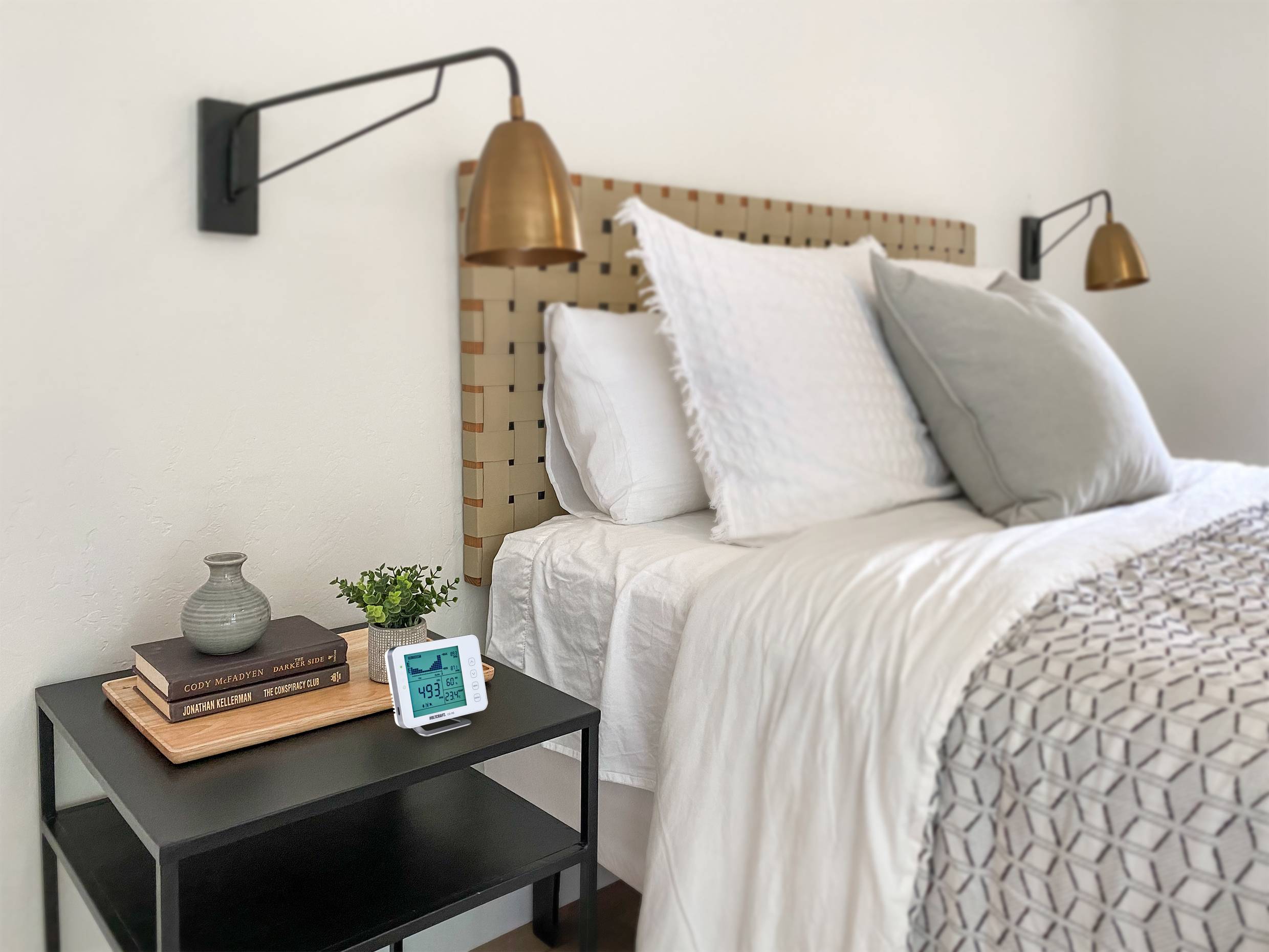 Une chambre simple avec un lit décoré, deux lampes murales et une table de chevet sur laquelle sont posés des livres, un vase et un réveil.