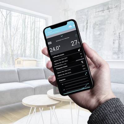Une personne tient un smartphone dans sa main, affichant une application météo. Température ambiante : 24°C, humidité : 27%. Salon en arrière-plan.