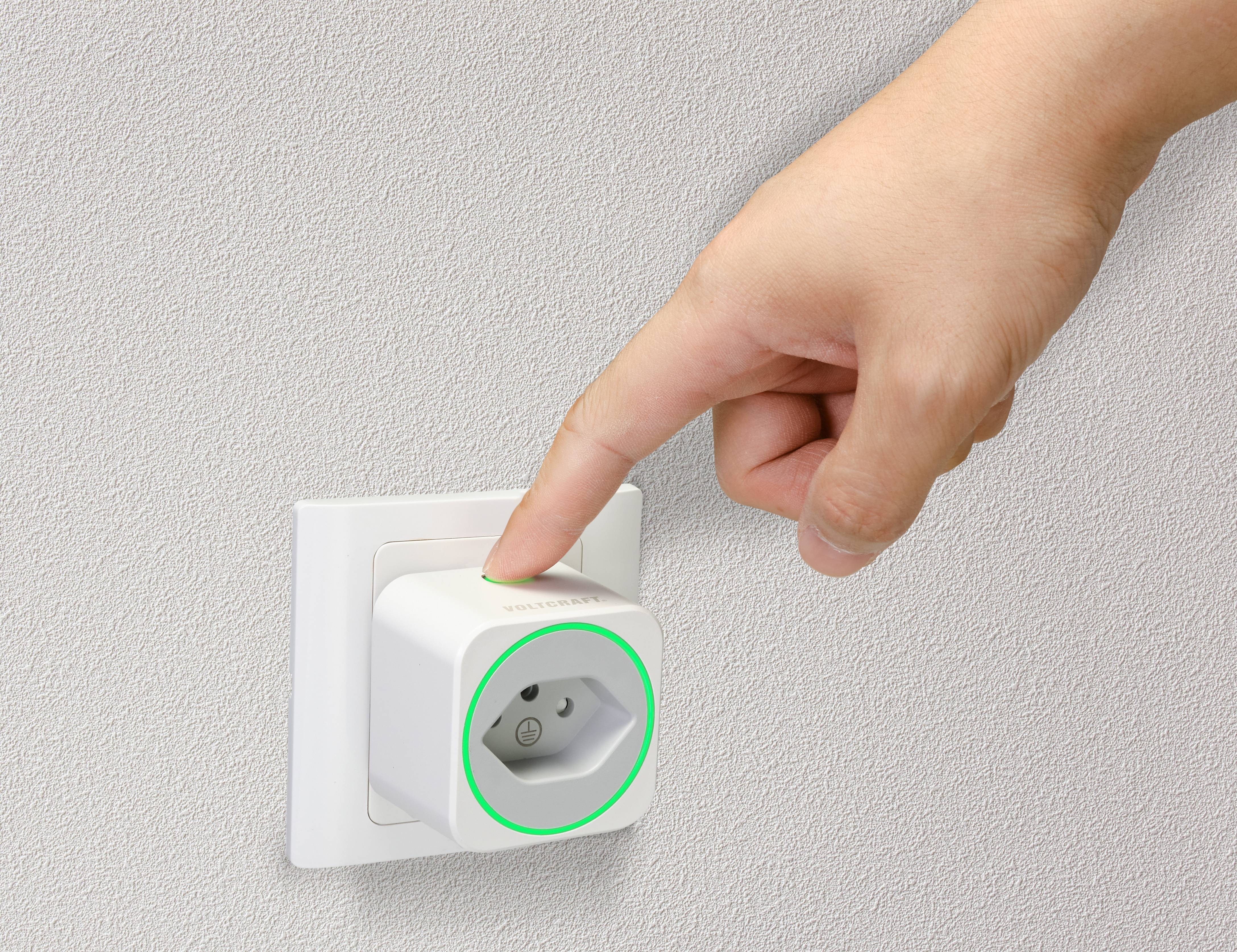 Une main appuie sur un adaptateur de prise intelligente connecté à une prise murale. Un anneau lumineux vert indique l'état de fonctionnement.