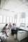 Trois personnes travaillent ensemble dans un bureau moderne. Elles sont assises à des bureaux avec des ordinateurs, entourées de grandes fenêtres.