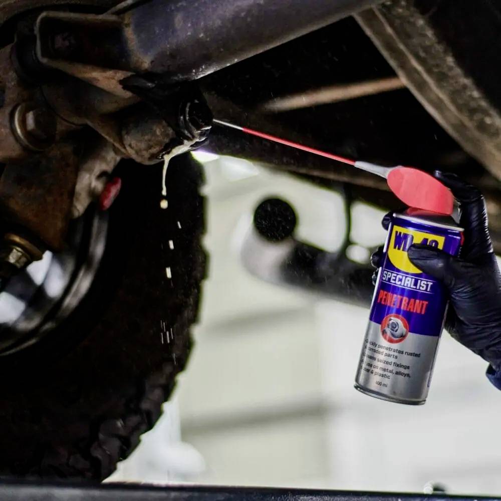 Une personne applique un lubrifiant sur une pièce automobile pour éliminer la rouille. Elle tient une bombe aérosol étiquetée « WD-40 Specialist ».