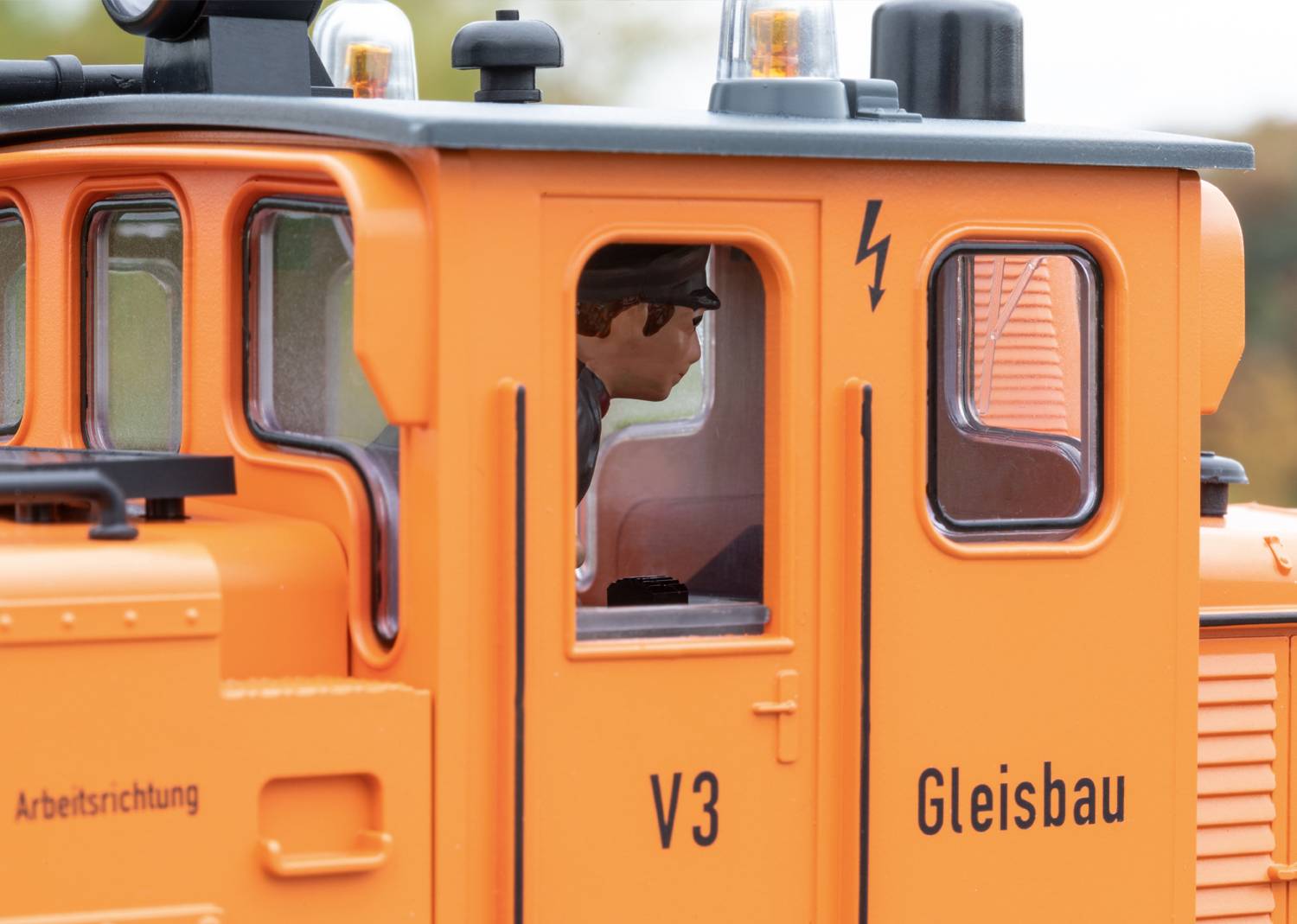 Tracteur de construction ferroviaire orange avec l'inscription « V3 », direction de travail marquée vers la gauche. Une personne est assise dans la cabine.