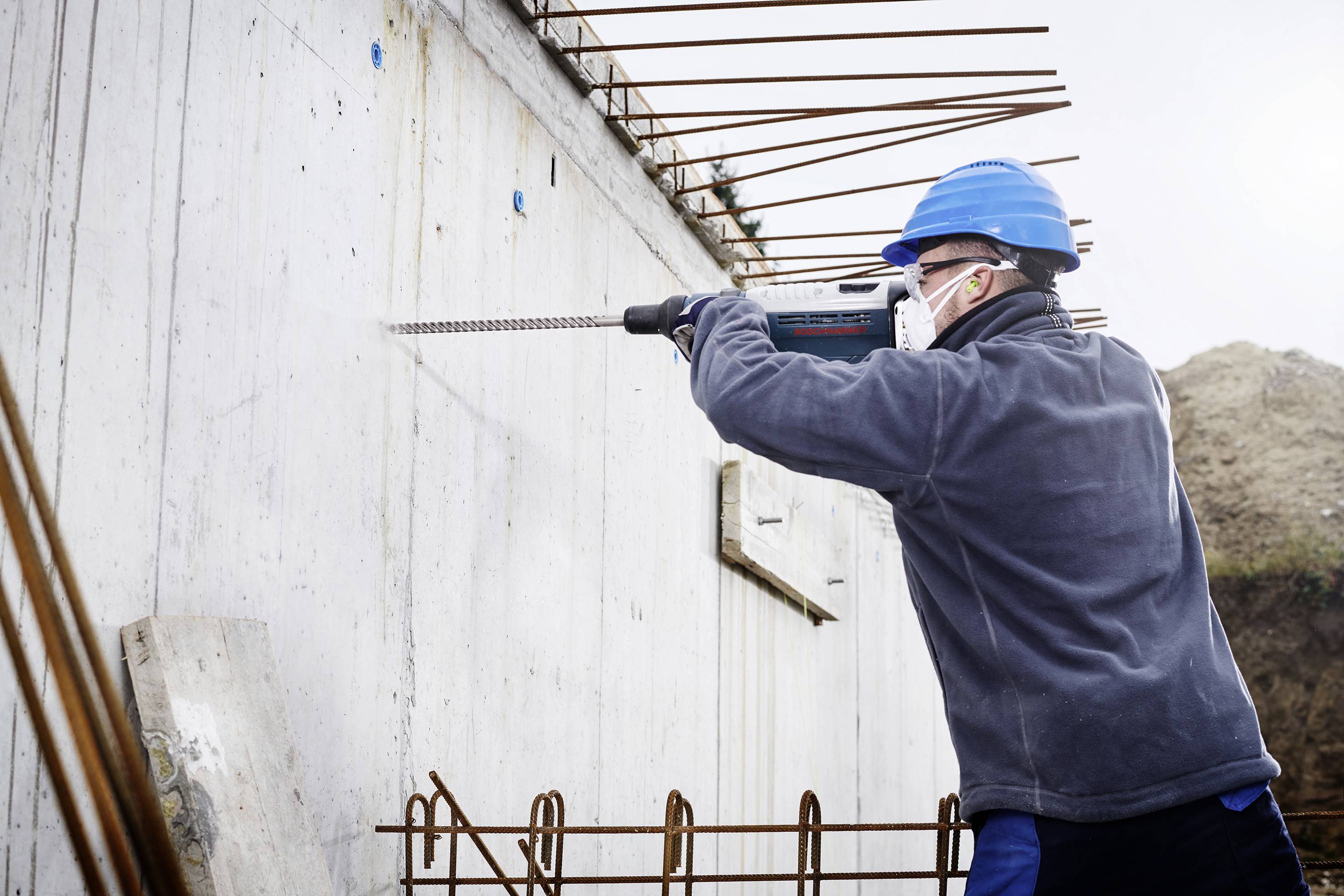Un ouvrier portant un casque de protection et un masque facial utilise une perceuse sur un mur en béton sur un chantier de construction.