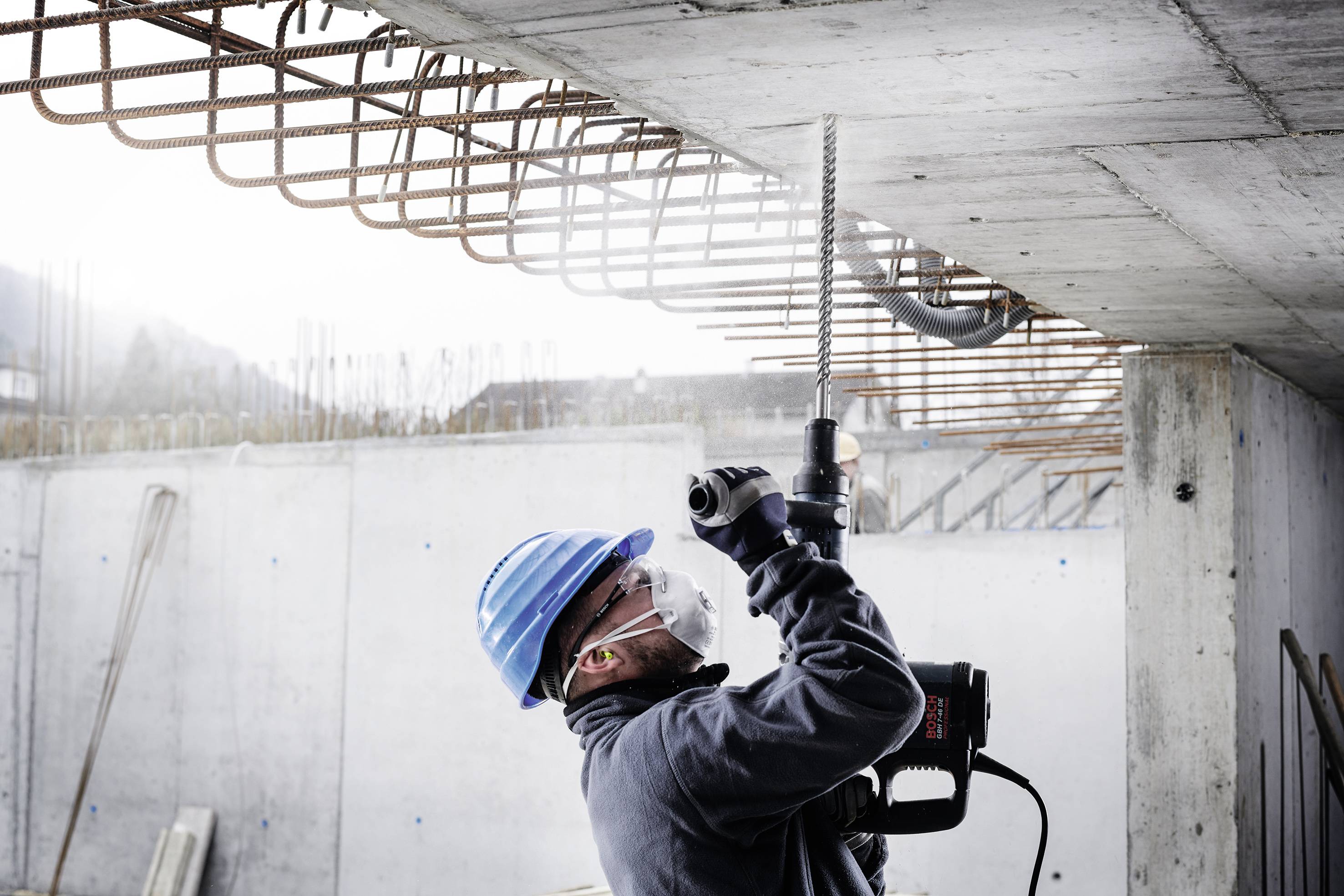 Un ouvrier porte un casque bleu et un masque alors qu'il travaille avec une perceuse sur un plafond en béton. Des poutres en acier sont visibles en arrière-plan.