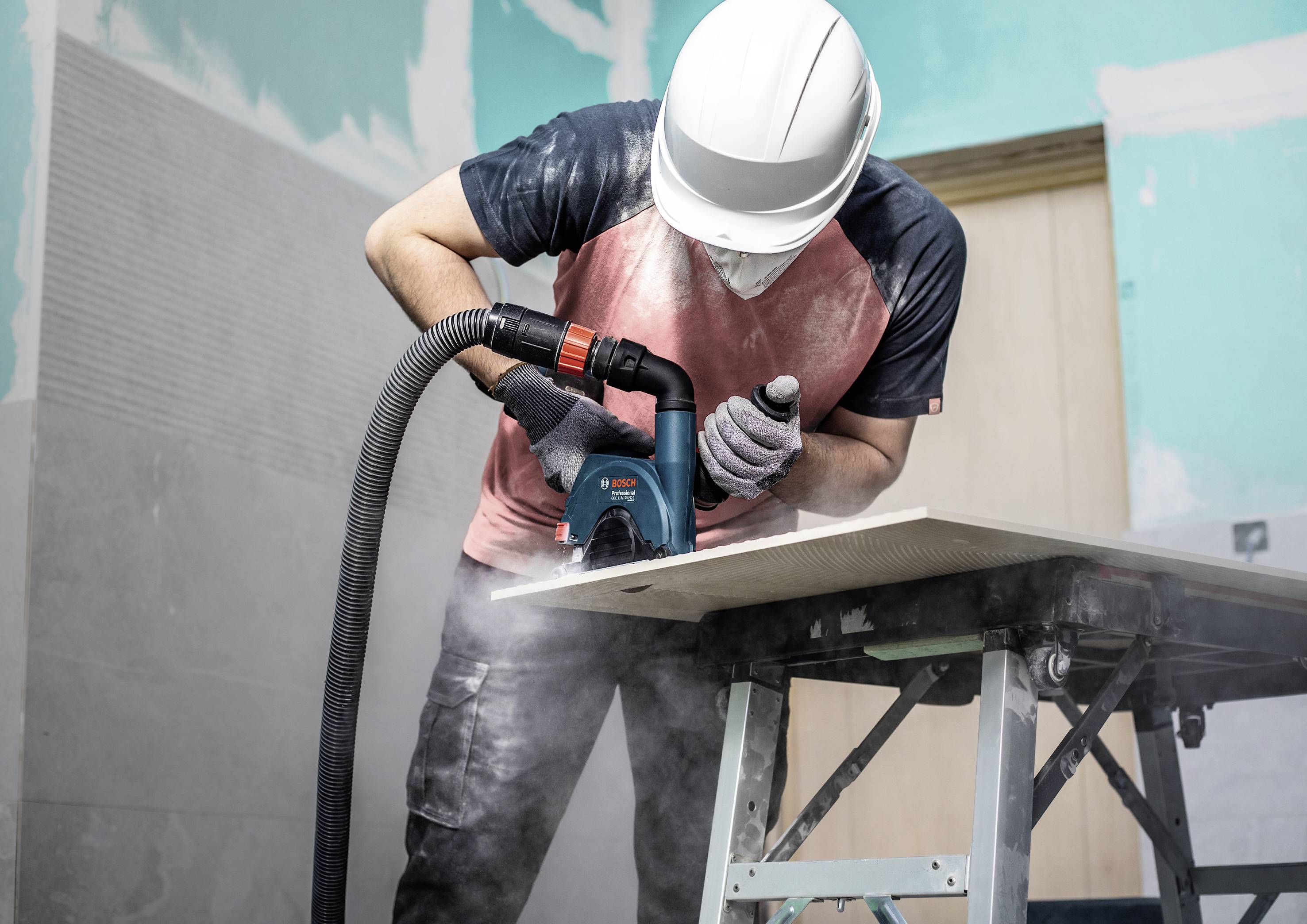 Un ouvrier porte un casque et un masque de protection, coupe une plaque avec une scie électrique, tandis que la poussière est aspirée.
