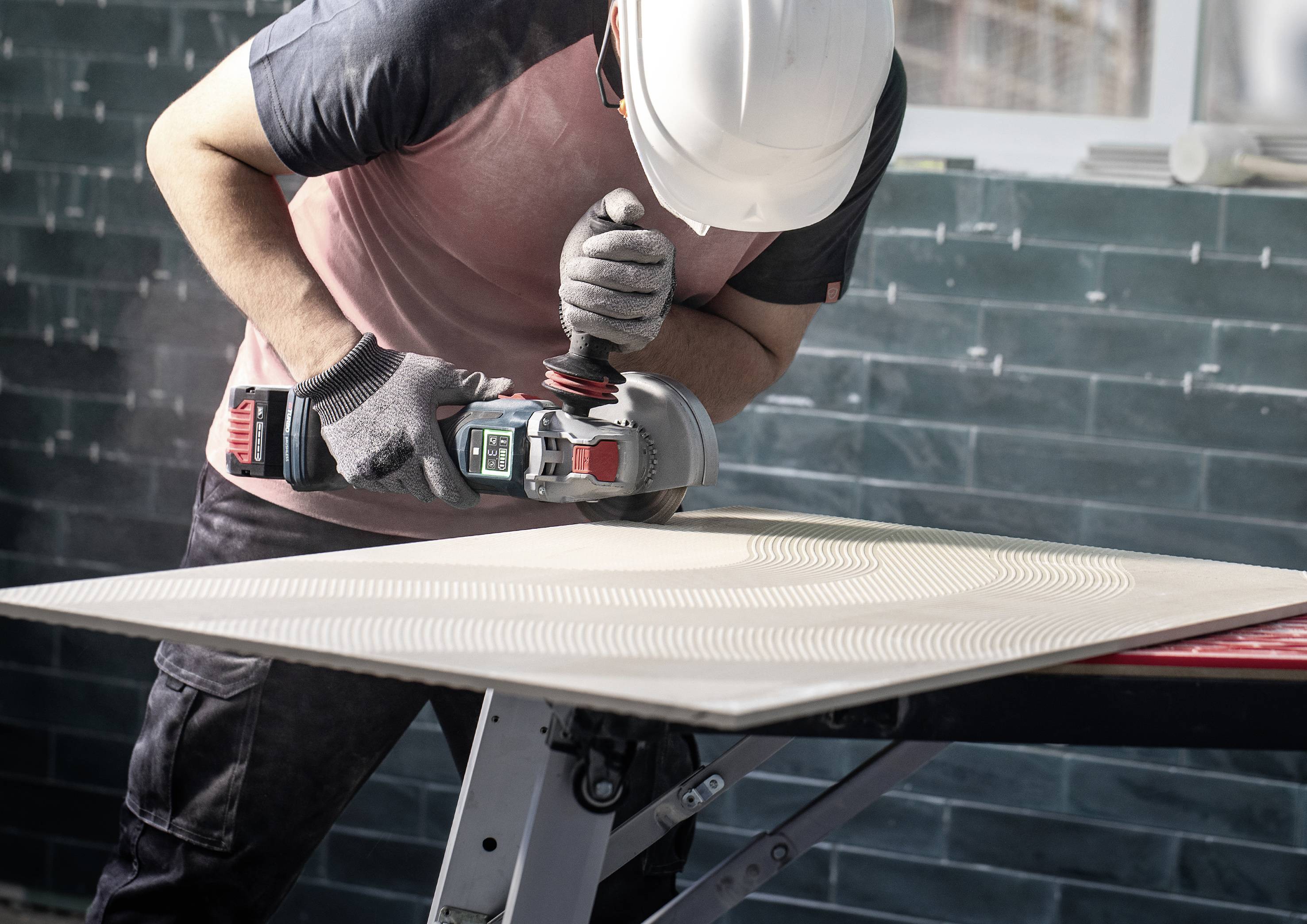 Un ouvrier portant un casque de sécurité ponce une grande planche de bois dans un atelier. Il utilise pour cela un meuleuse d'angle électrique.