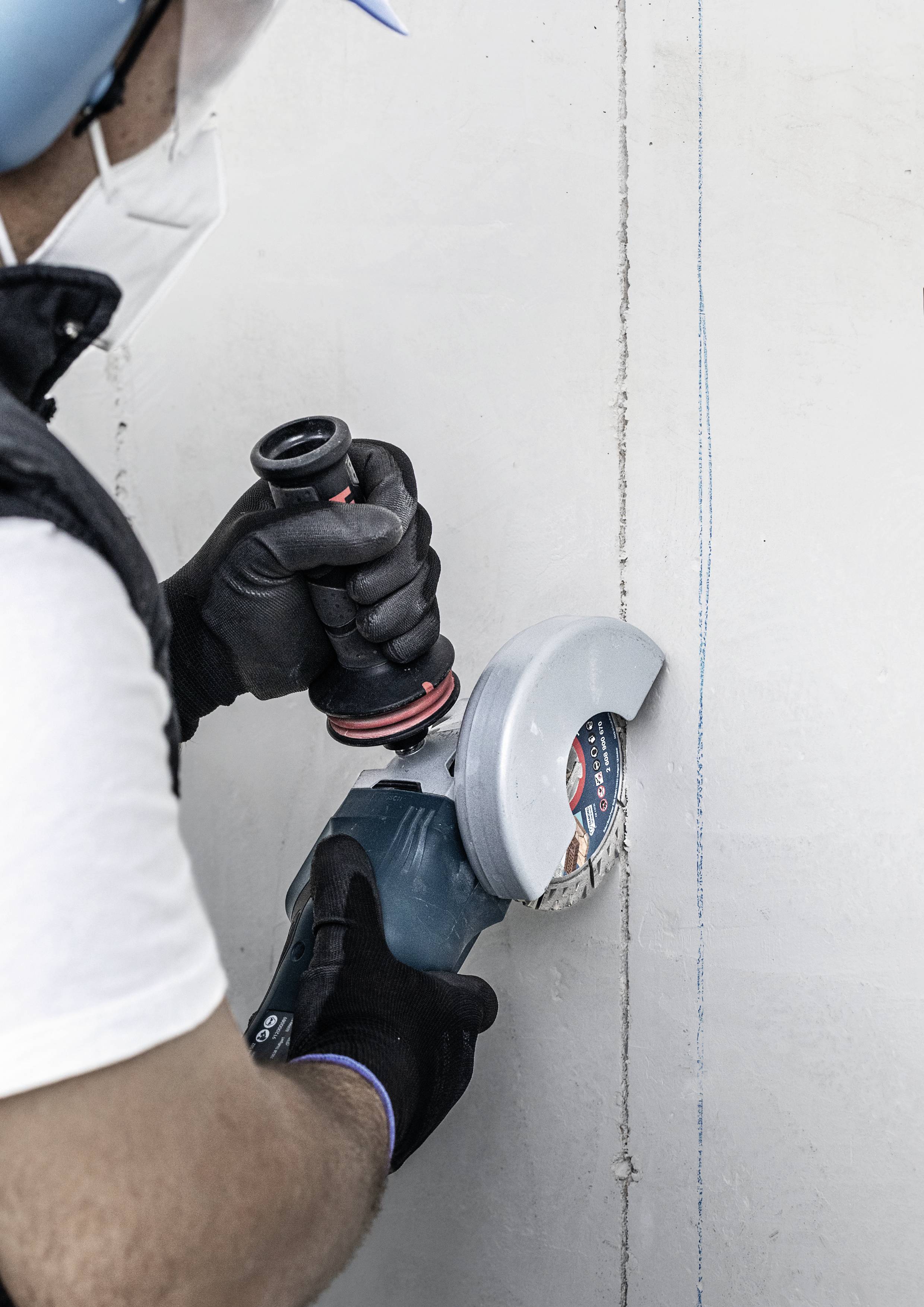 Une personne portant un masque de protection et un casque meule un mur en béton à l'aide d'une meuleuse d'angle.