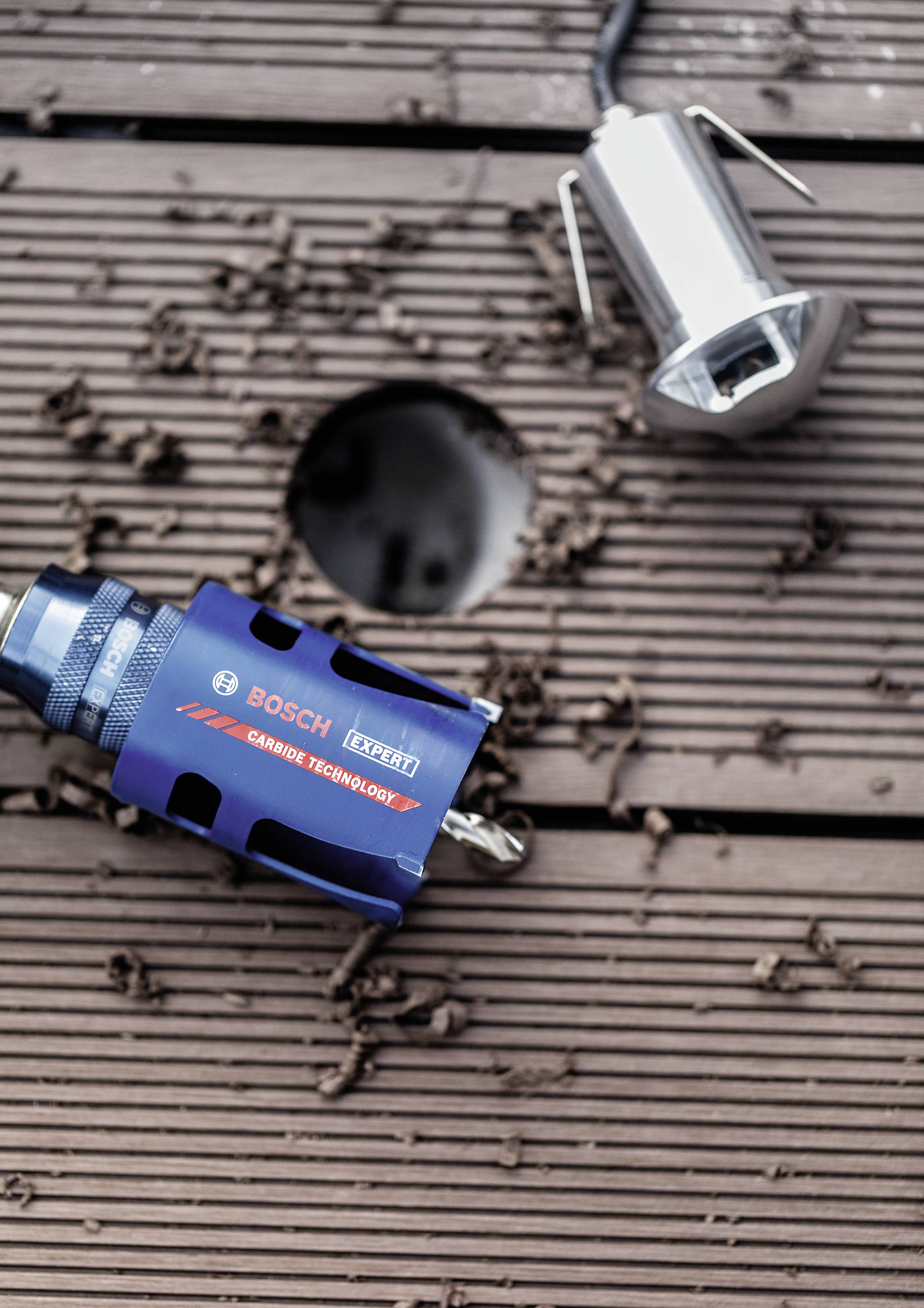 Un embout de perceuse bleu portant la marque 'Bosch' est posé à côté d'un trou et d'une lumière métallique sur une surface en bois. Des copeaux de bois sont dispersés çà et là.