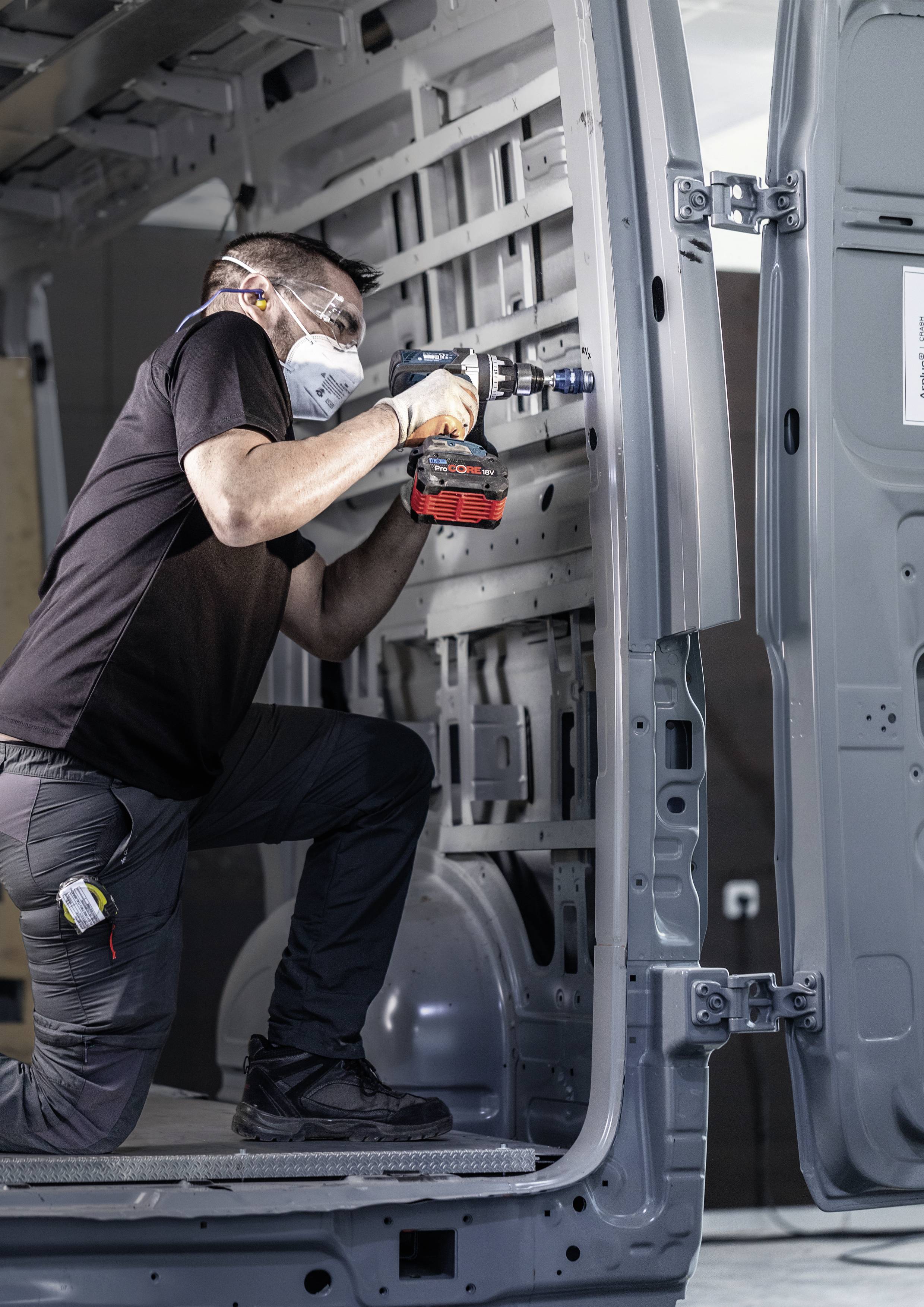 Un artisan en t-shirt noir et masque de protection perce l'intérieur d'un fourgon. Il travaille avec concentration sur l'aménagement du véhicule.
