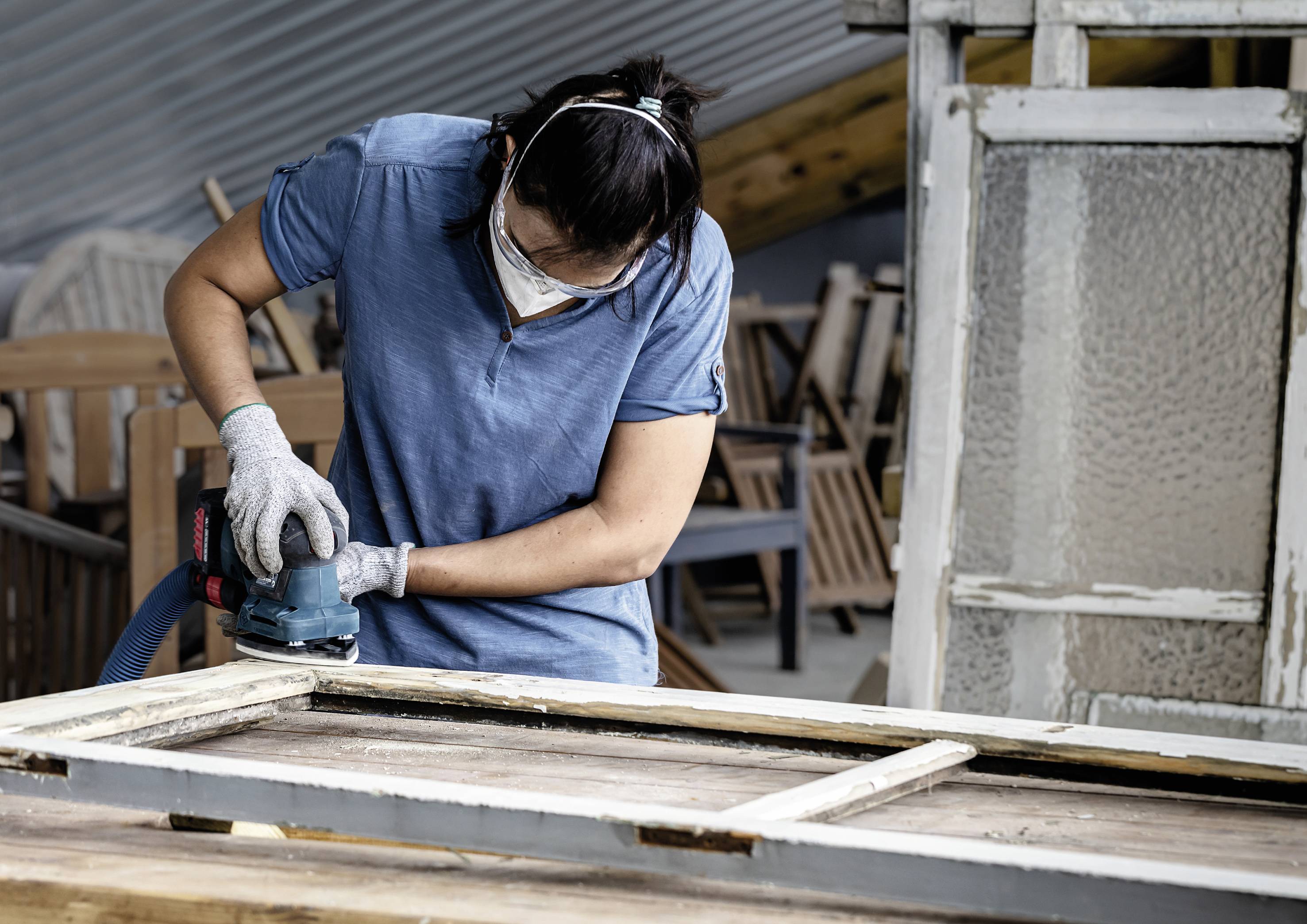 Une personne ponce délicatement une ancienne fenêtre dans un atelier. Des lunettes de protection et un masque sont portés. L'arrière-plan montre des meubles.