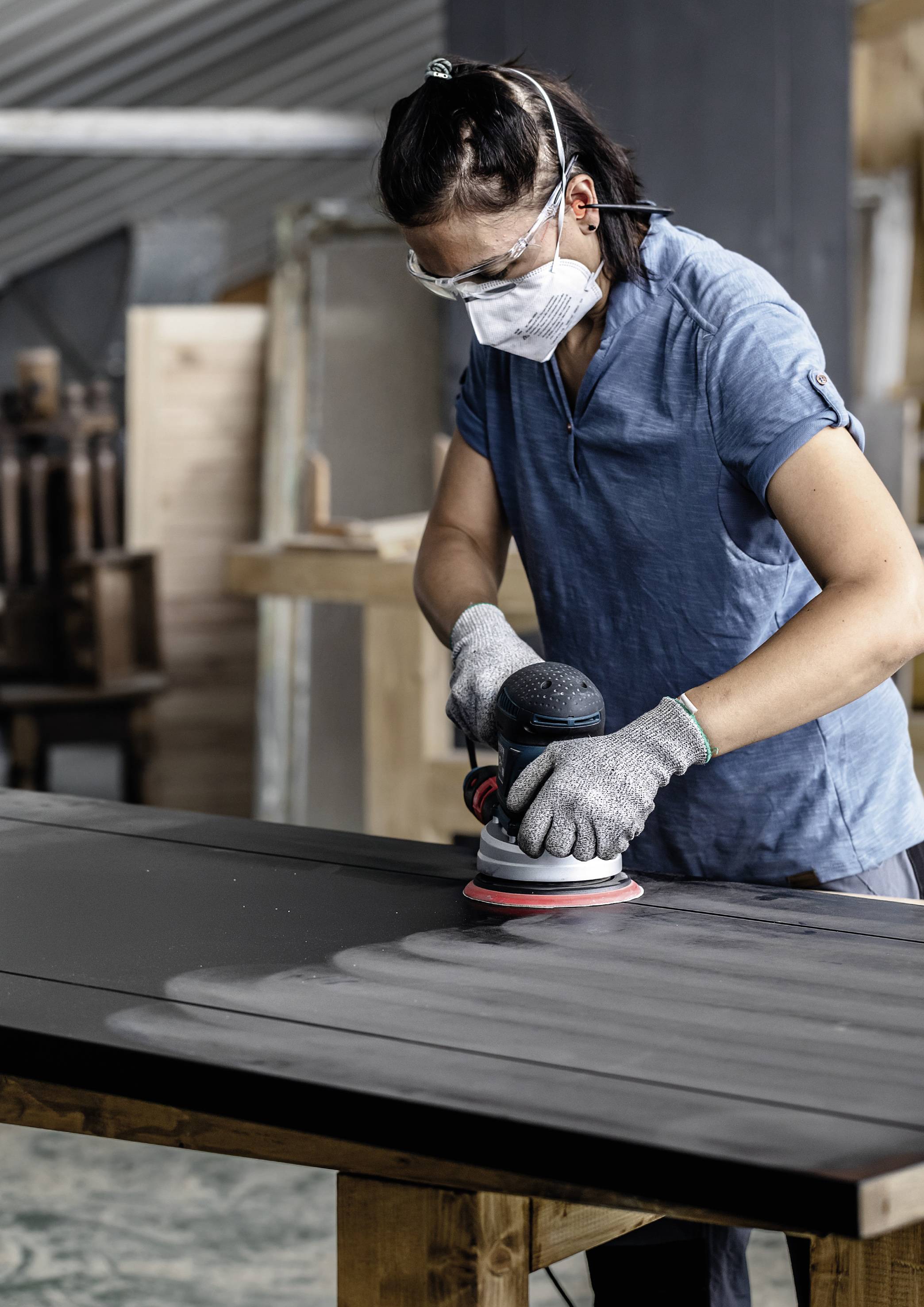 Une personne porte des lunettes de protection et un masque et ponce un plateau de table en bois dans un atelier.