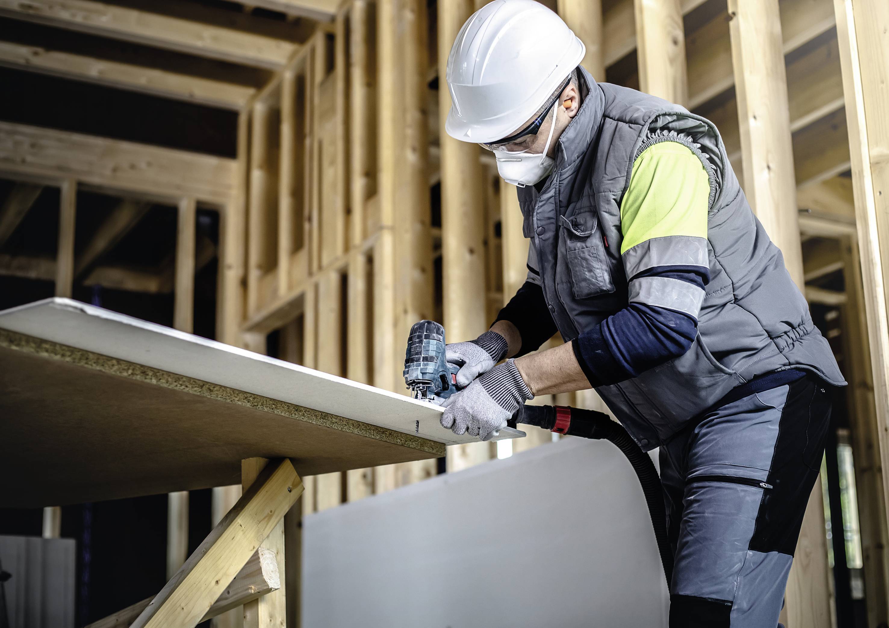 Un ouvrier portant un casque de chantier et un masque utilise une perceuse pour fixer des panneaux de bois dans un bâtiment.