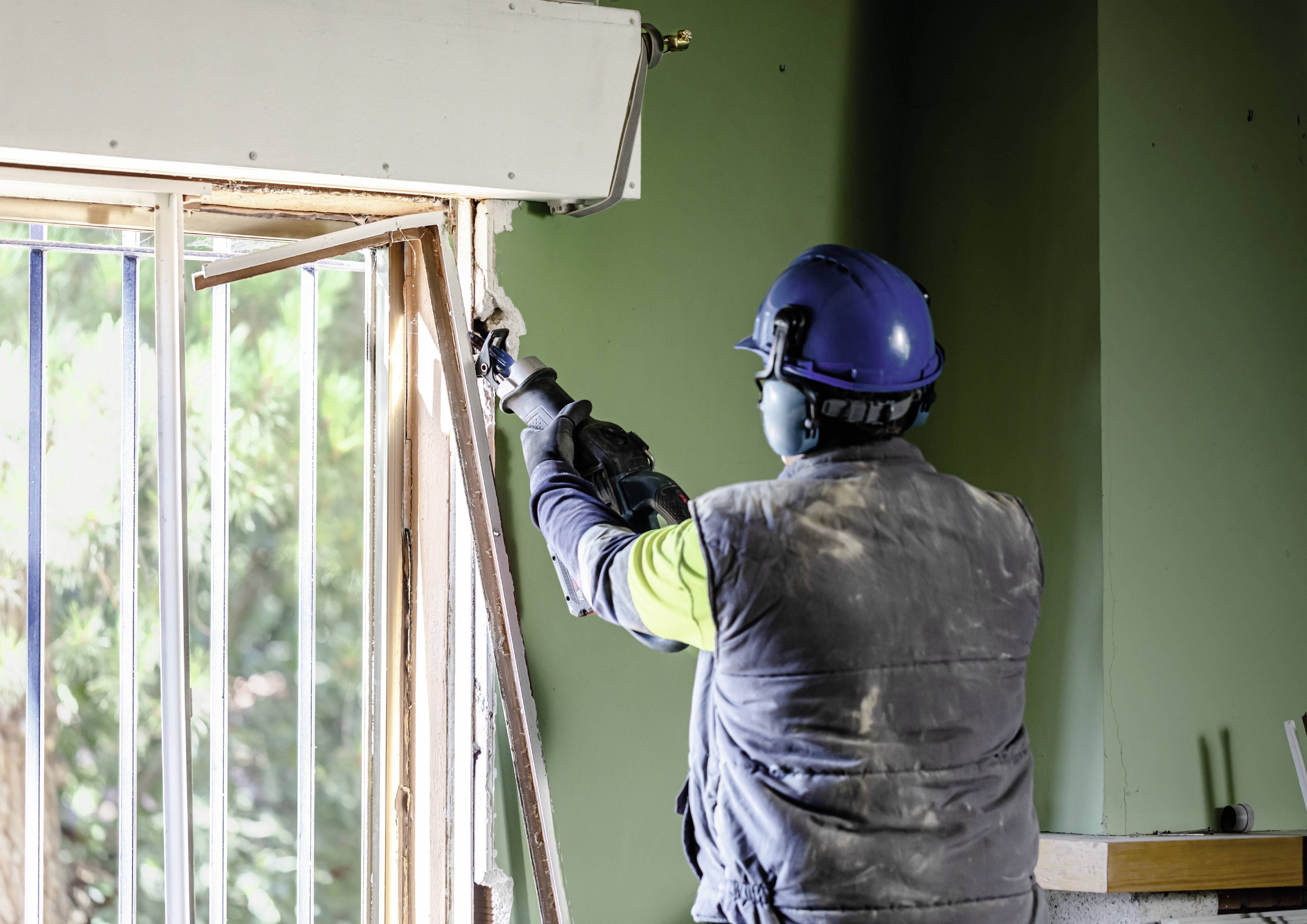 Un artisan portant un équipement de protection et un casque travaille avec un outil électrique sur un cadre de fenêtre dans une pièce verte.