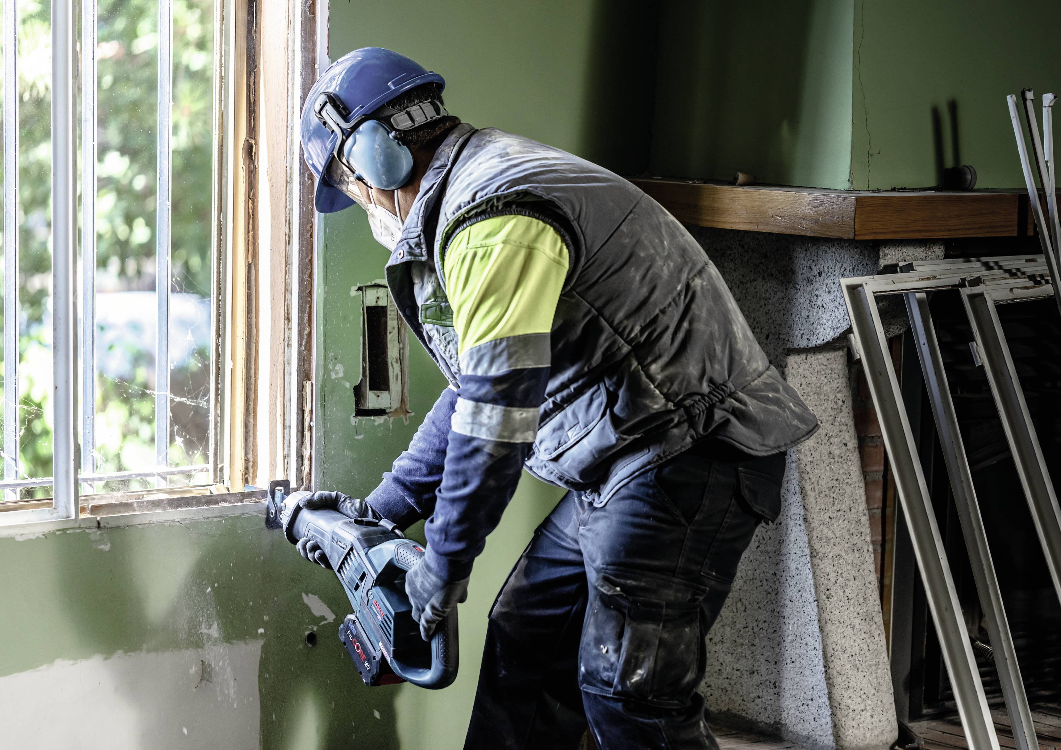 Un ouvrier portant un équipement de protection utilise un outil électrique pour travailler une fenêtre dans une pièce aux murs verts.