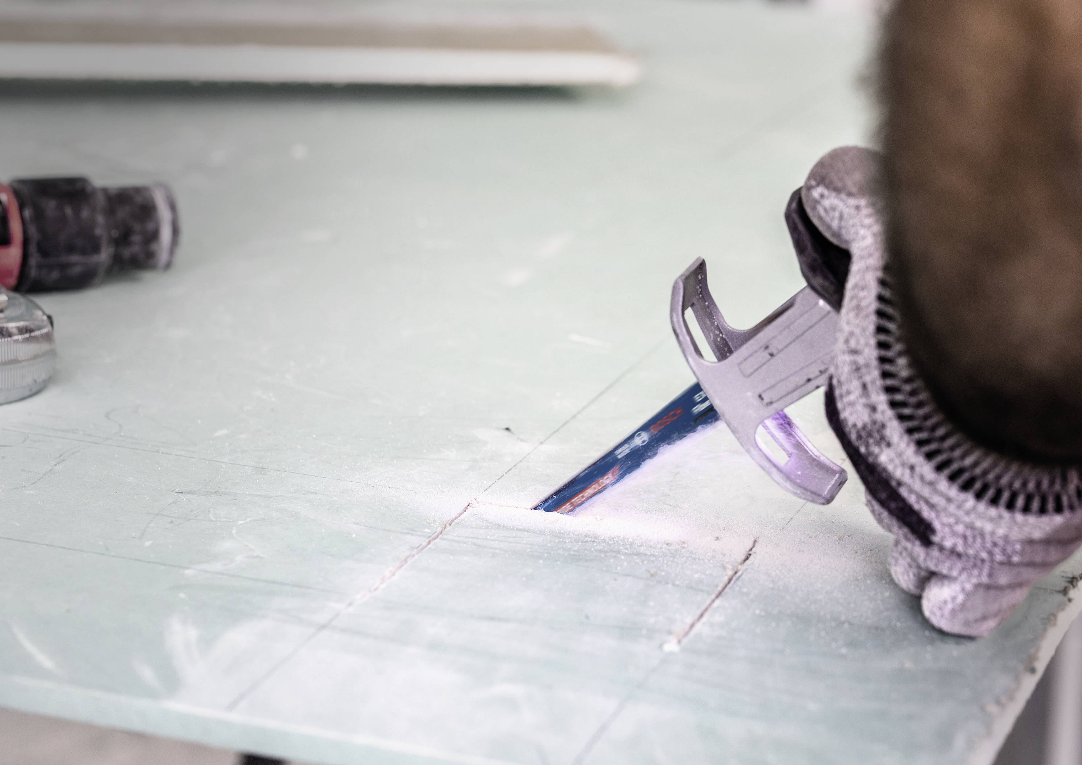 Une personne coupe une plaque de plâtre verte à l'aide d'une scie. La personne porte des gants de travail, et des copeaux de bois sont visibles.