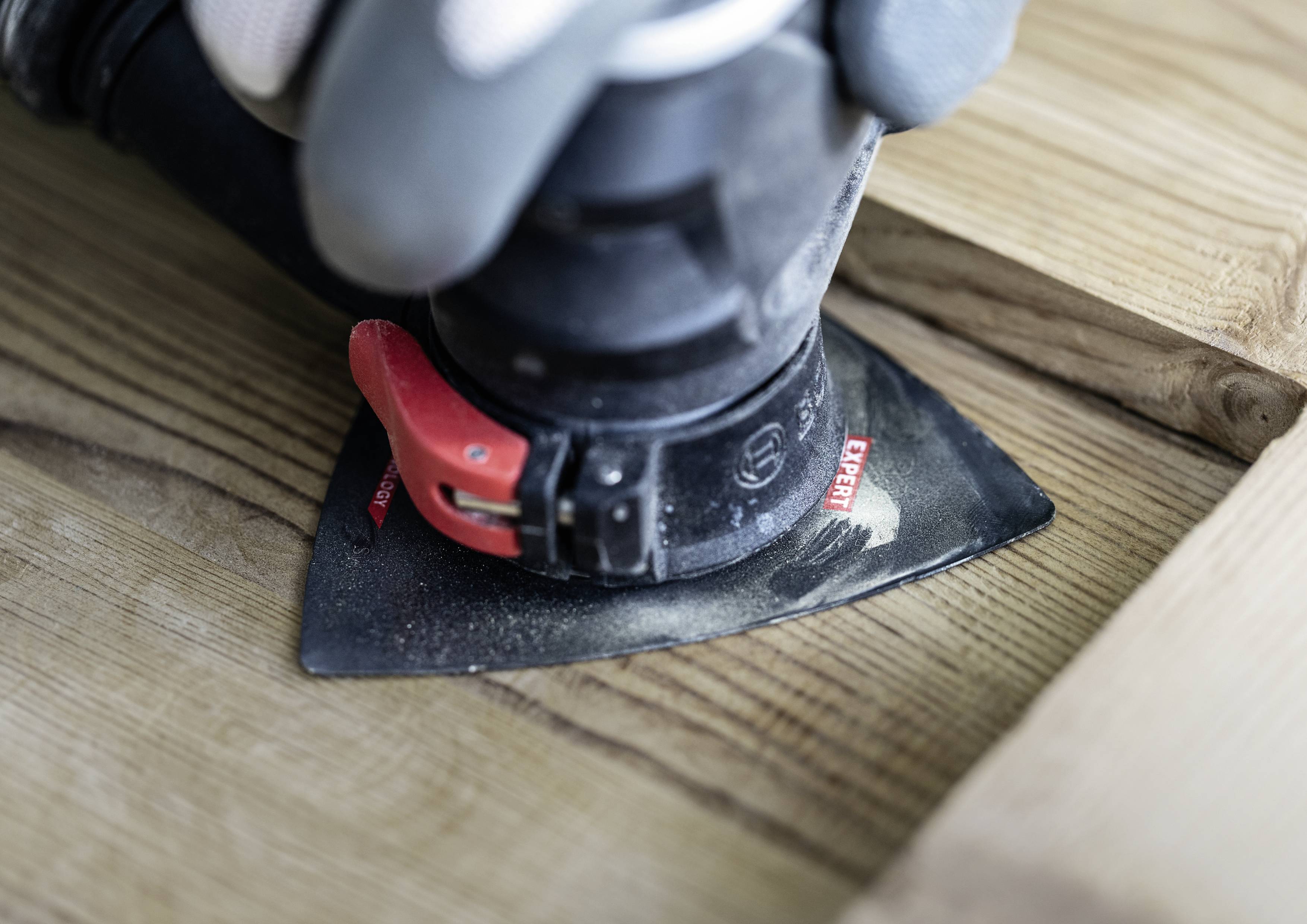 Une main gantée tient un appareil de ponçage et travaille une surface en bois. Le papier de verre a une forme triangulaire.