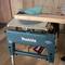 Une scie circulaire manuelle bleue posée sur un support de table Makita vert. Une personne est en train de régler la machine. Des copeaux de bois sont visibles en dessous.