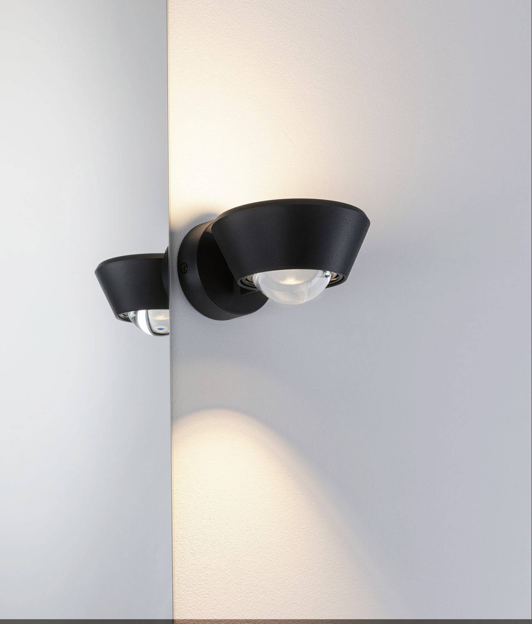 Une lampe murale moderne au boîtier noir projette une lumière chaleureuse sur un mur gris clair.