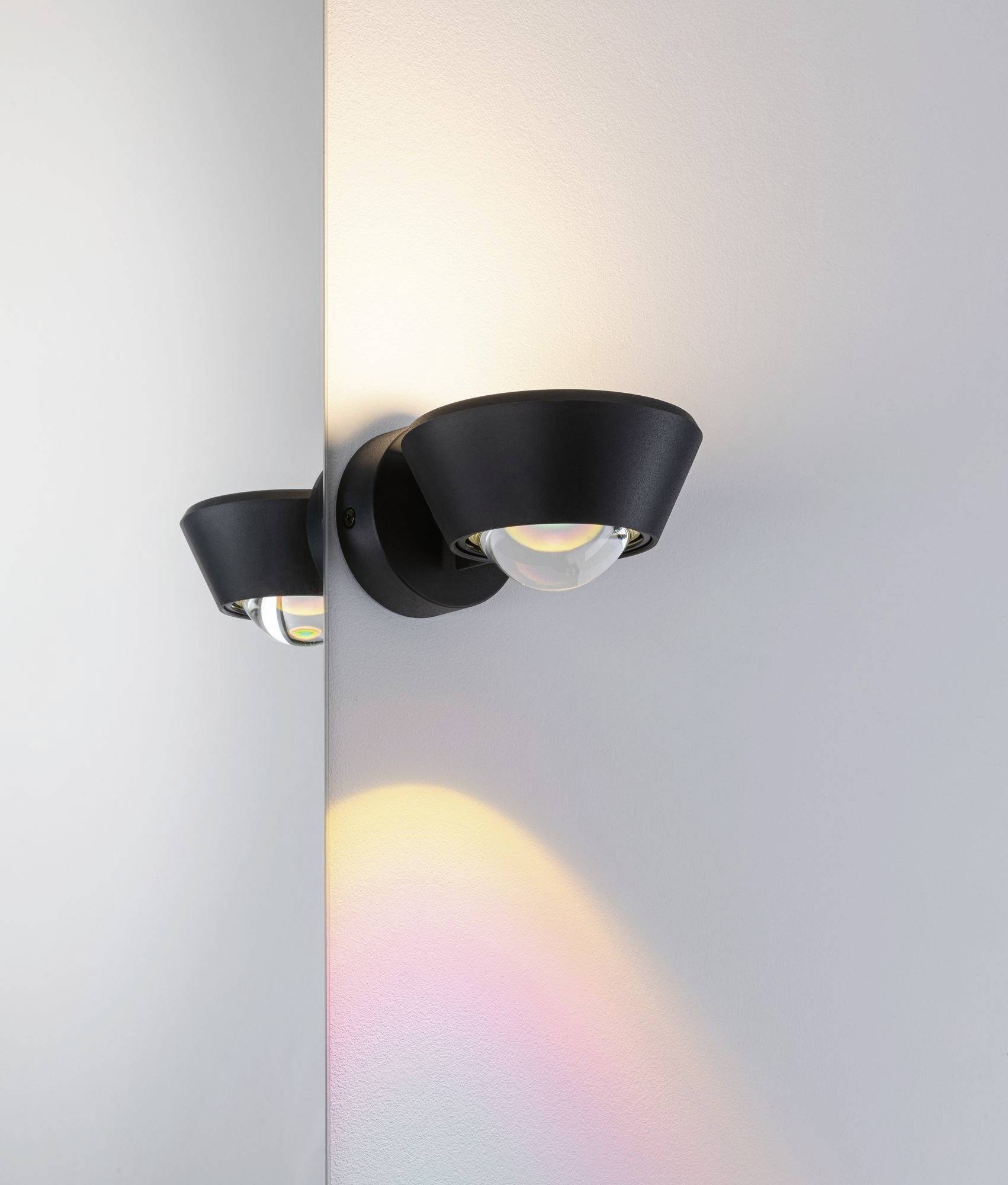 Un applique murale moderne, noir, sur un mur blanc, diffuse une lumière douce et colorée.