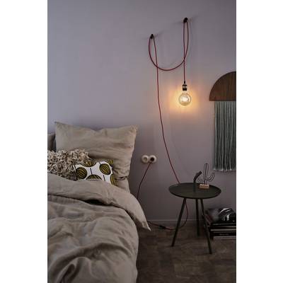 Chambre chaleureuse avec un lit décoré de coussins. À gauche, sur le mur, une ampoule suspendue. Table de chevet avec une décoration de cactus en dessous.