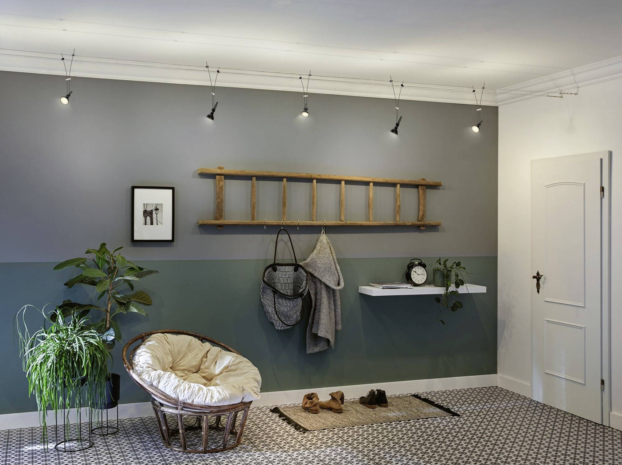 Un salon aux murs bleu-gris, avec une échelle en bois appuyée contre le mur, un fauteuil en osier garni de coussins, des plantes et une étagère sur laquelle est posée une horloge.