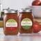 Deux bocaux de sauce tomate sur une table, chacun avec une étiquette rouge et verte. À l'arrière-plan, une tomate et des ustensiles de cuisine.