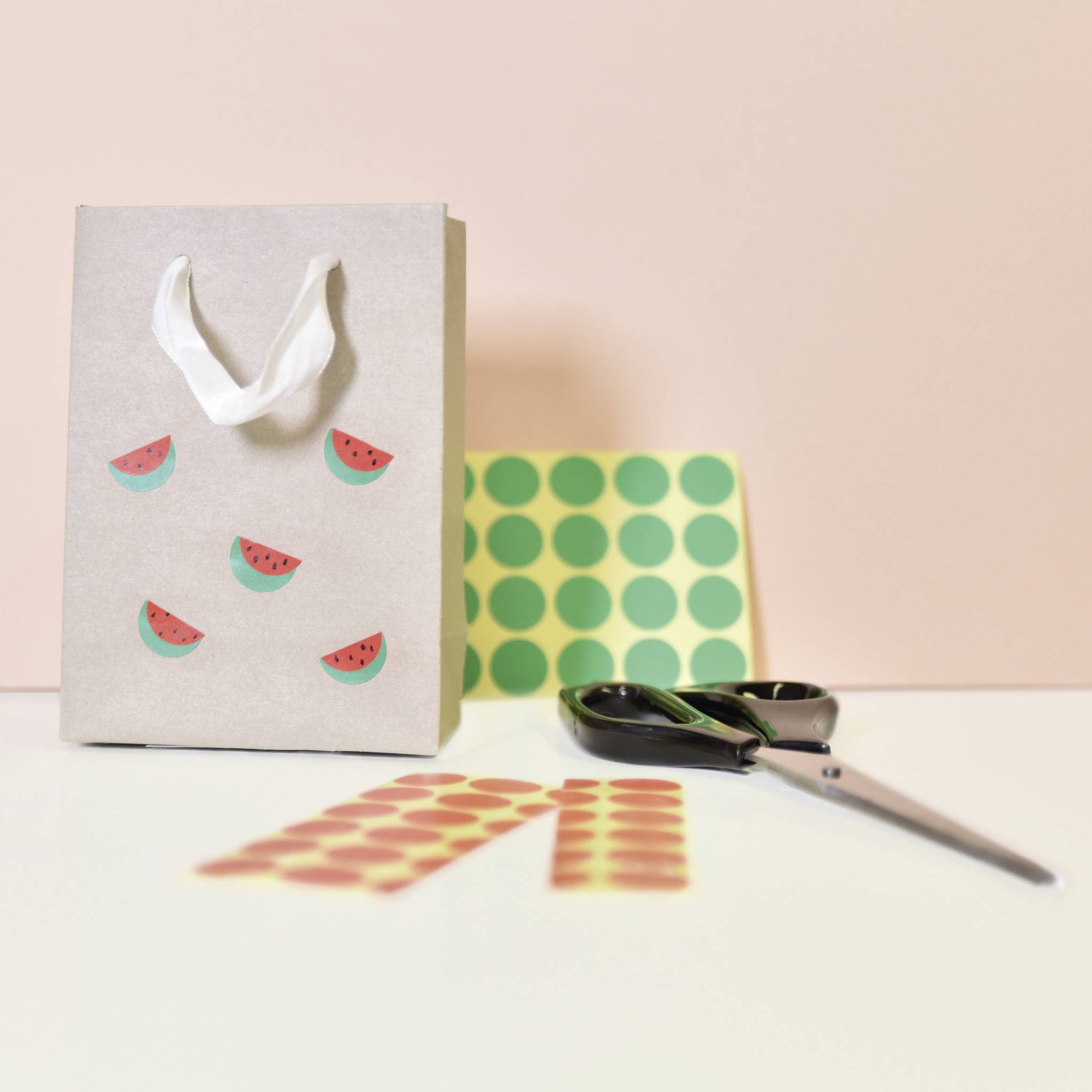 Un sac cadeau brun avec des autocollants de pastèques, à côté d'une paire de ciseaux et de points adhésifs colorés sur une table blanche.