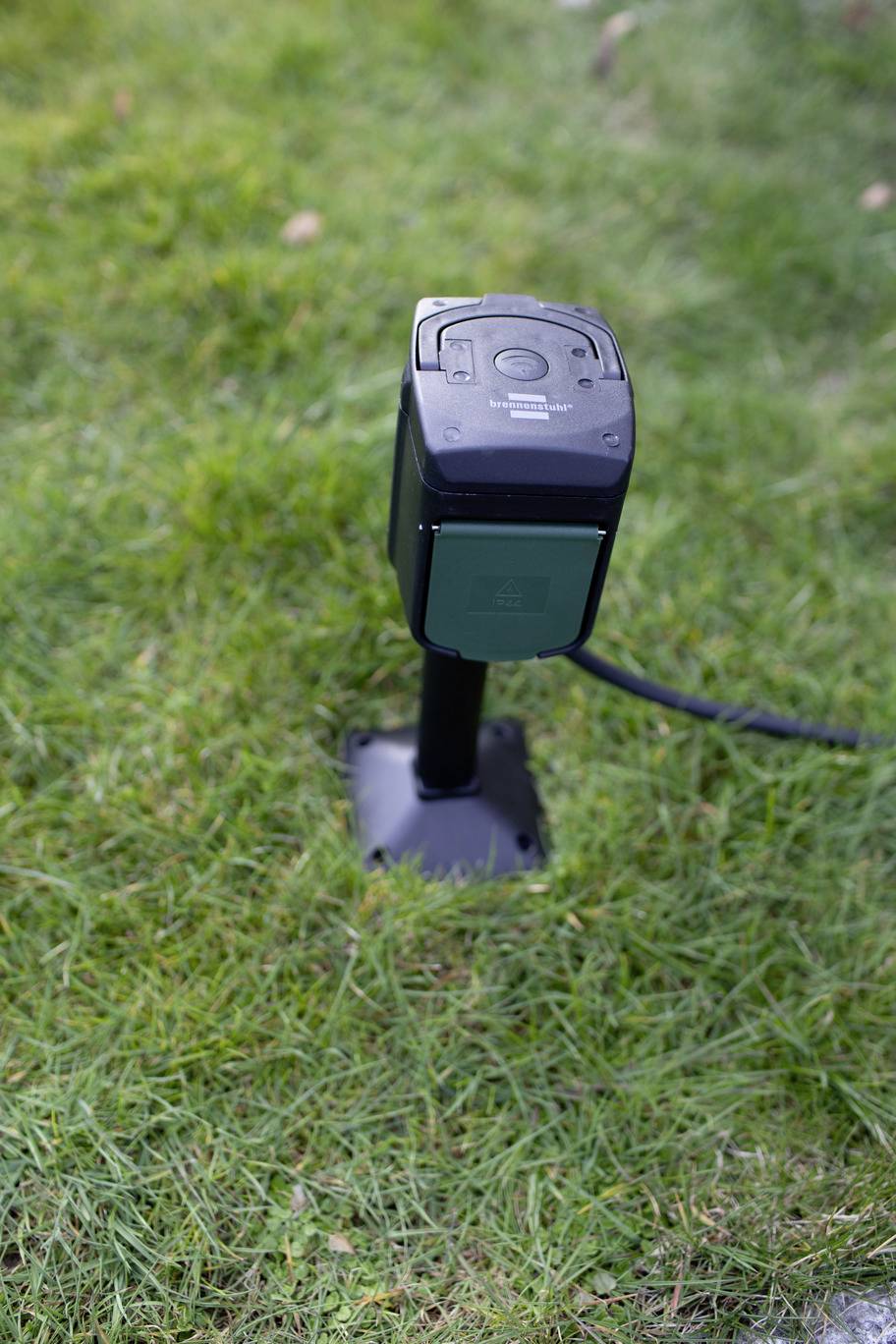 Une prise électrique noire est posée sur un gazon vert. Un câble s'en échappe. Elle est conçue pour une utilisation en extérieur.