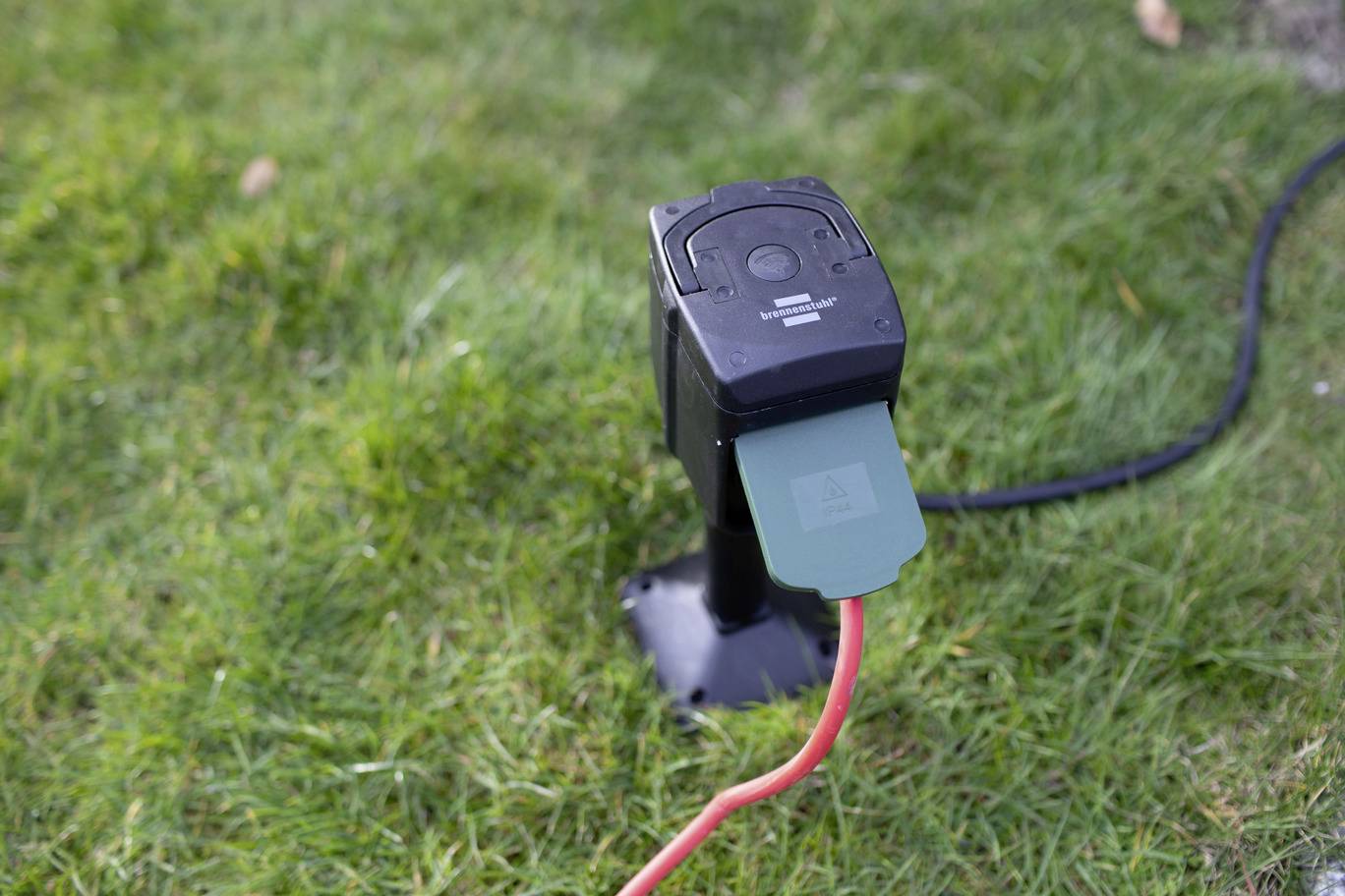 Prise électrique sur un poteau planté dans la pelouse, reliée à un câble rouge, entourée d'herbe.