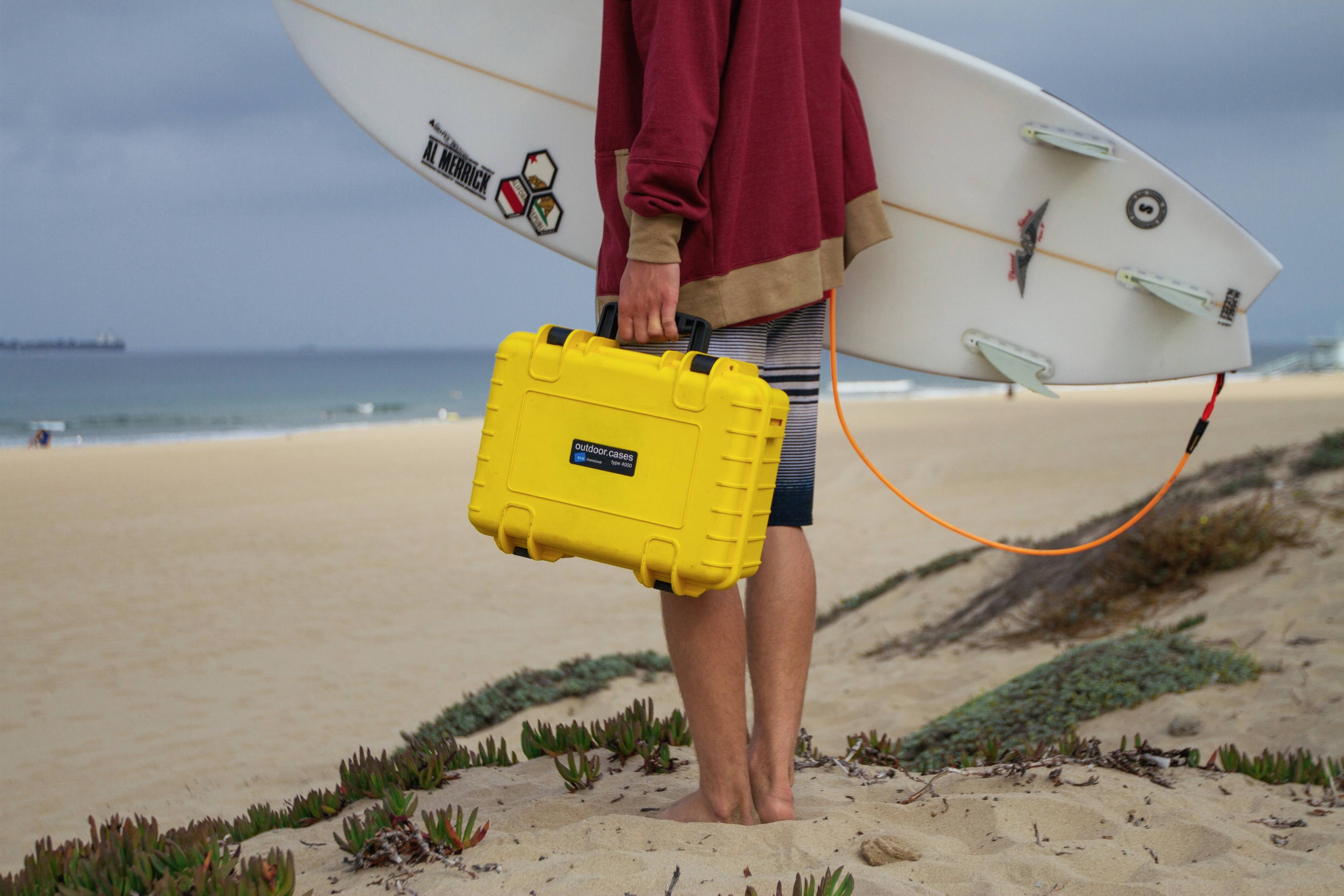 Une personne se tient sur la plage, tenant une planche de surf et une valise jaune à la main, prête à surfer.