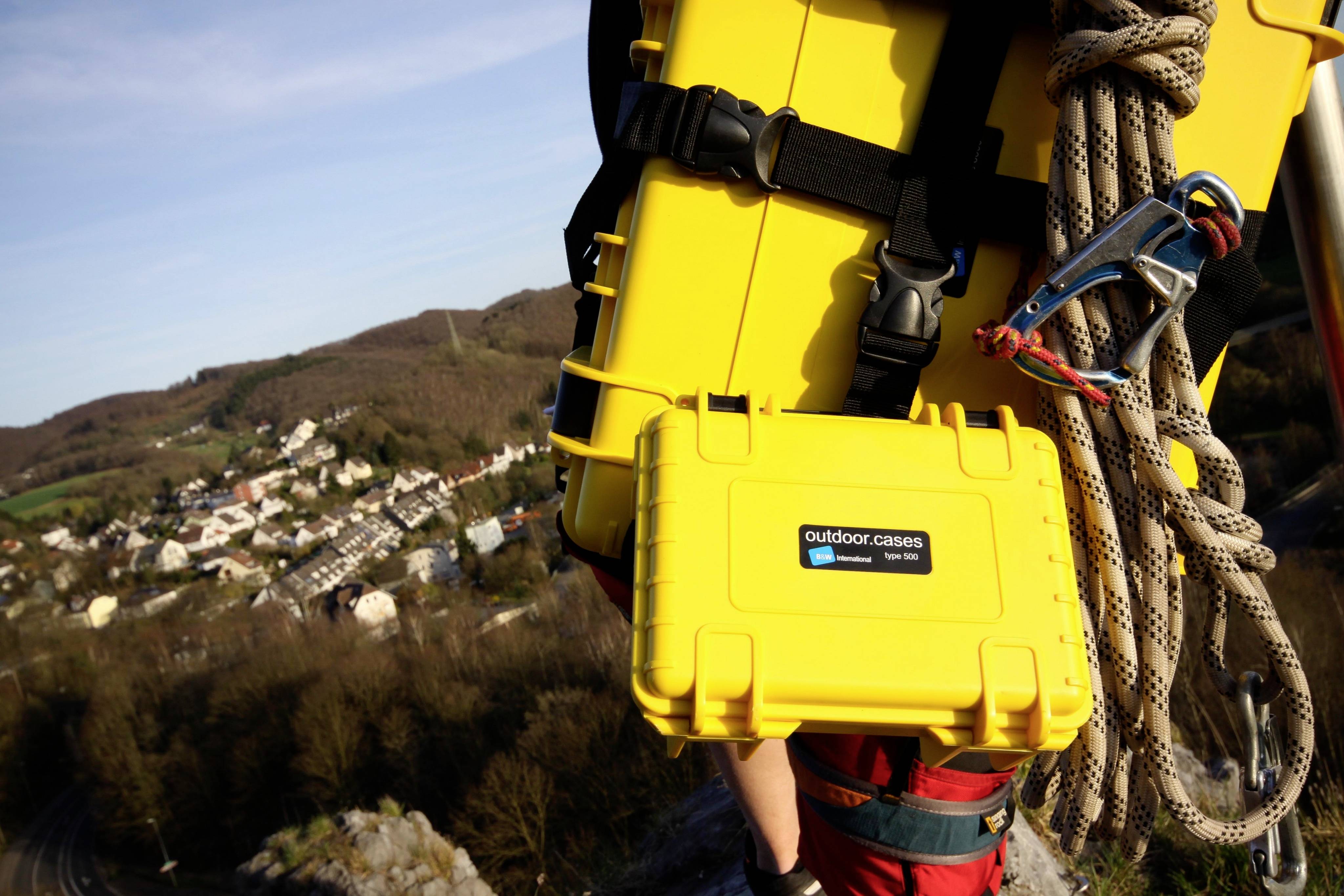 Une personne porte deux valises rigides jaunes et une corde sur le dos lors de l'escalade. À l'arrière-plan, un paysage avec des maisons est visible.